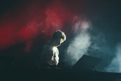 一位身穿白衬衫的音乐家站在舞台上，身后的烟雾中透出红白相间的灯光